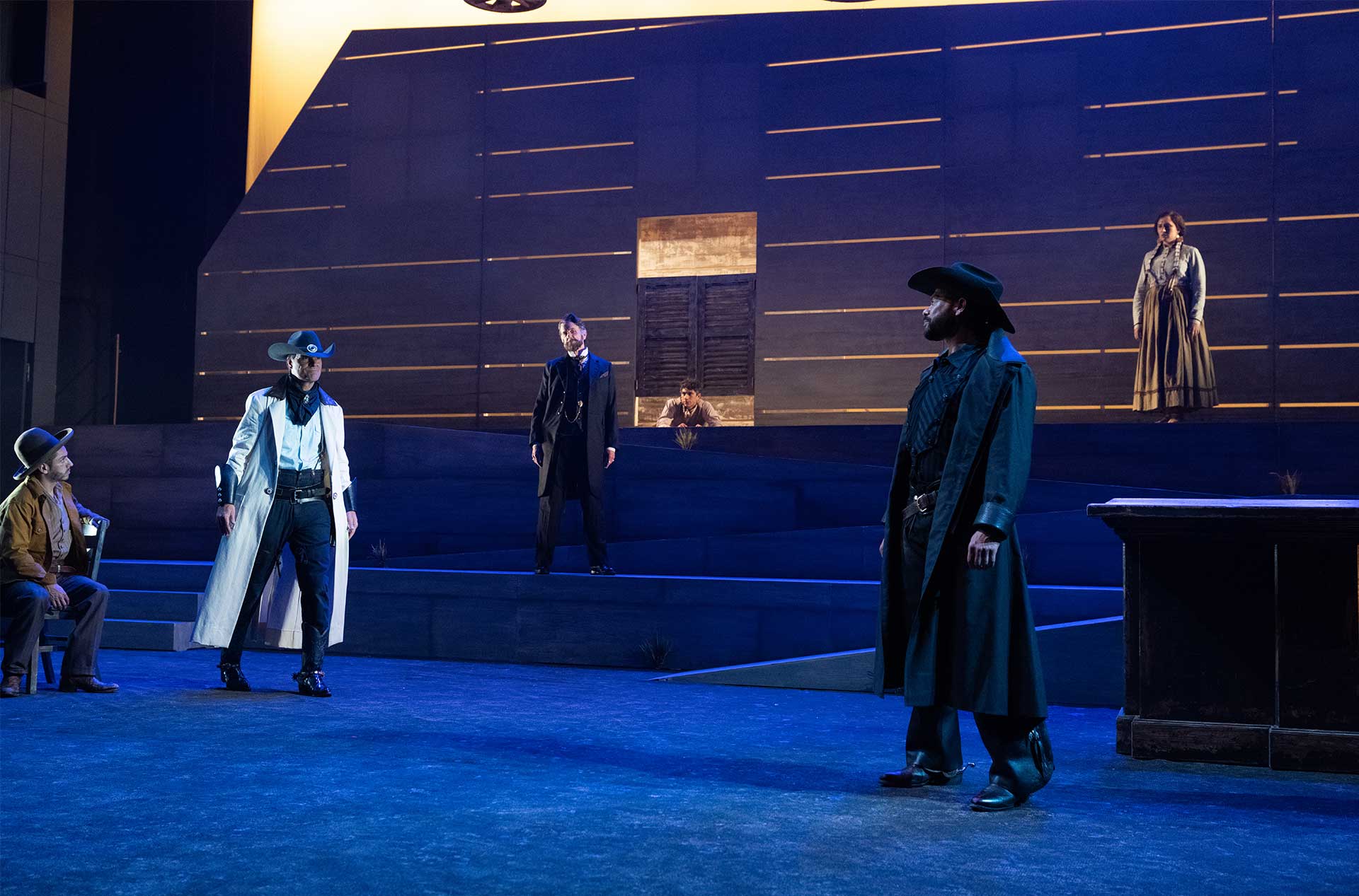 A standoff scene with multiple performers in western attire spaced across a layered stage, with dramatic lighting and a central figure in a white coat.
