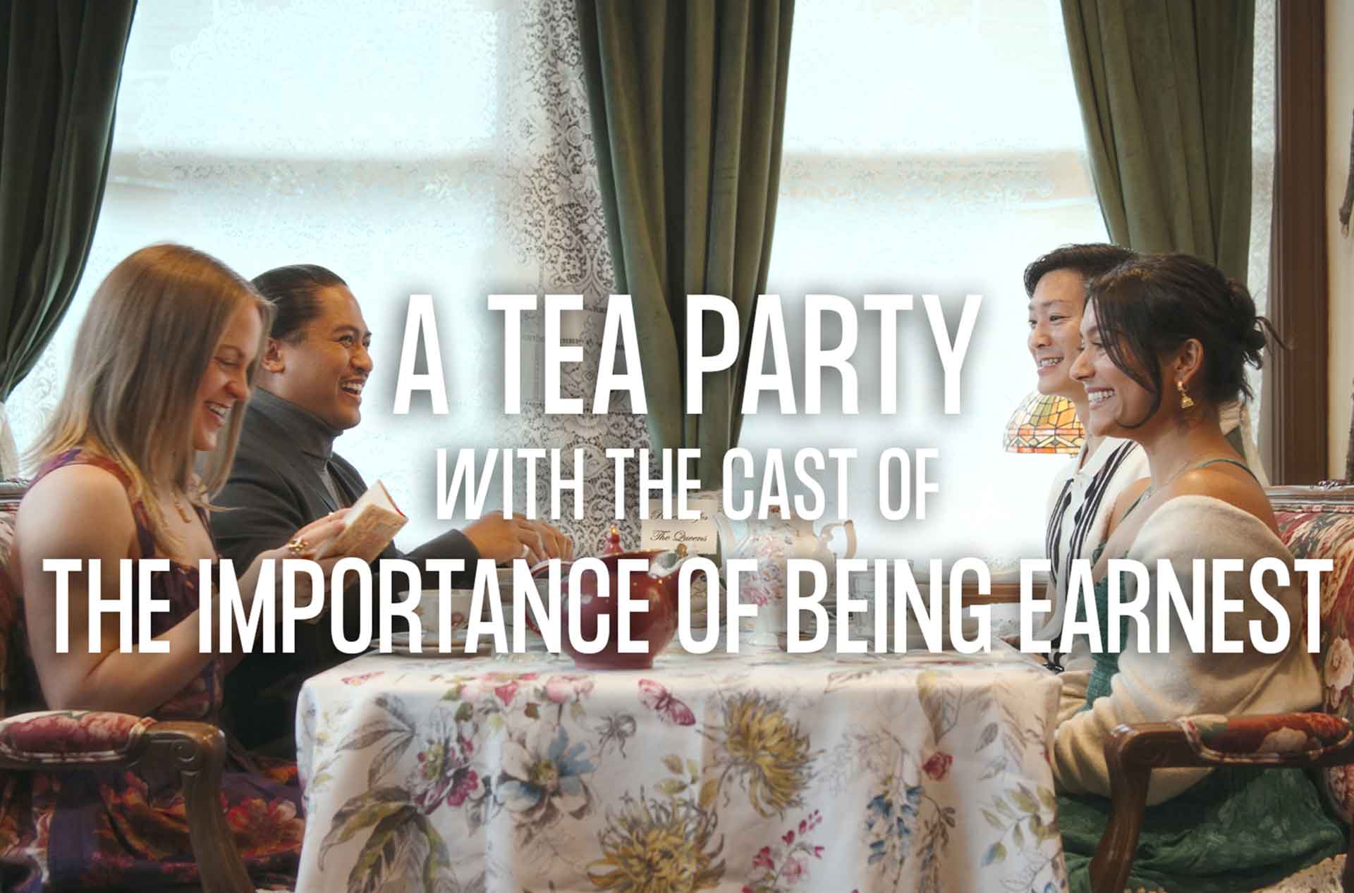 Four cast members smile and laugh while gathered around a floral tablecloth for a tea party, promoting "The Importance of Being Earnest."