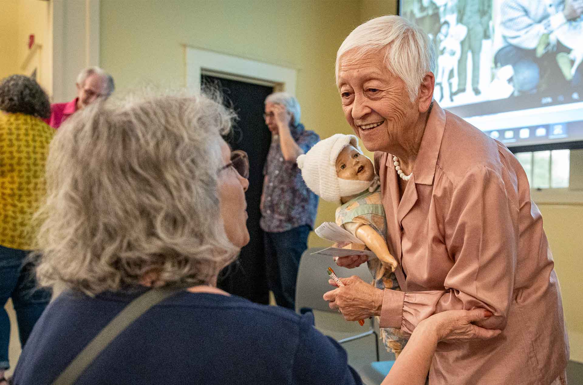 An older individual with short hair smiles while holding a vintage doll and speaking with another person in a group setting. People are gathered in the background with a projected image on the wall.