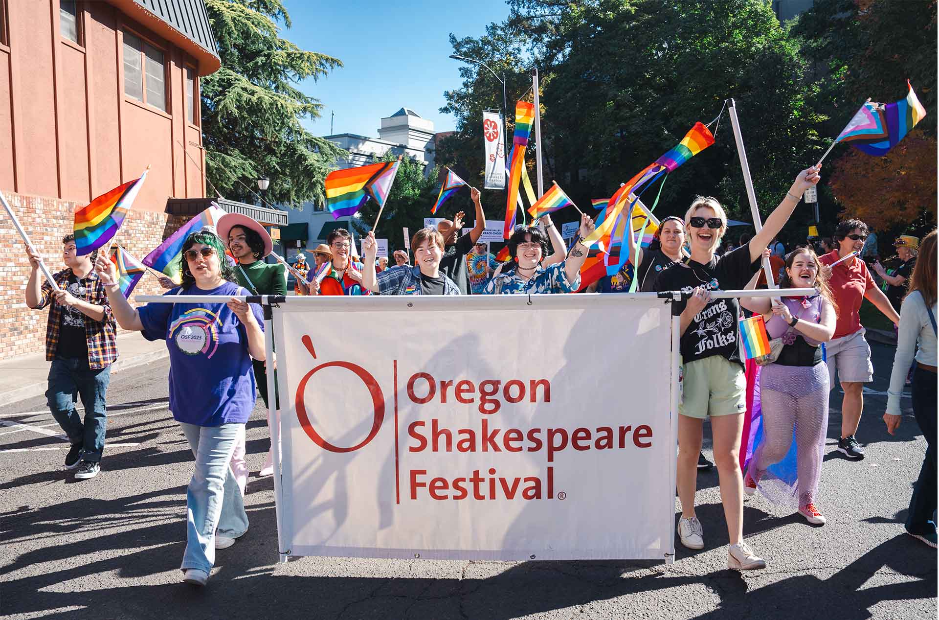A group of people marches in a parade holding a banner that reads "Oregon Shakespeare Festival," waving pride flags. The crowd appears joyful as they walk down the street.