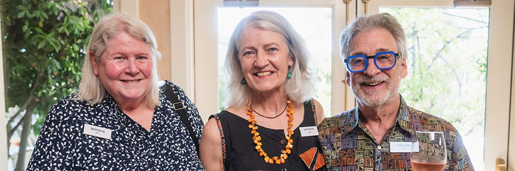 Three people smile and pose for a photo indoors, wearing name tags and holding drinks during a social gathering.