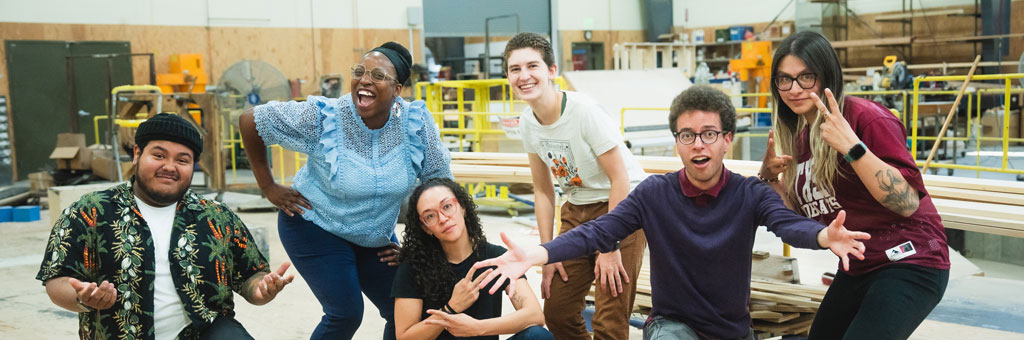 A diverse group of people poses cheerfully in a workshop space with wood and construction materials in the background.