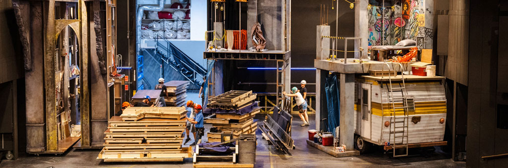 A colorful and detailed theater set features stacked wooden pallets, construction elements, and a camper with ladders and scaffolding.
