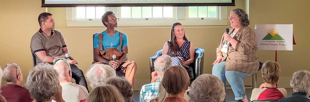 Four panelists sit on stage in front of an audience, each holding microphones. A sign behind them displays &quot;Ashland Sunrise Project,&quot; indicating the event theme.