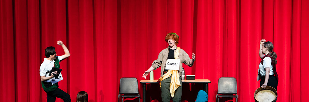 Theatrical performance depicting Julius Caesar’s assassination; student actors on stage with labeled signs, dramatic poses, and Roman-themed costumes.
