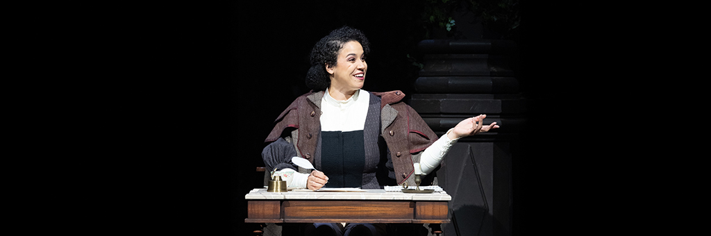 Person in a brown and gray period costume sits at a wooden desk with an inkwell and papers, smiling and gesturing to the right. Dark background with a stone pillar suggests an indoor setting.