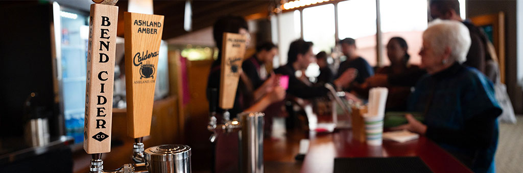 Close-up of a cider tap labeled "Bend Cider Co" alongside another tap labeled "Ashland Amber Caldera Brewing Company," with people blurred in the background a concession stand. The scene conveys a social atmosphere with patrons interacting while ordering drinks.