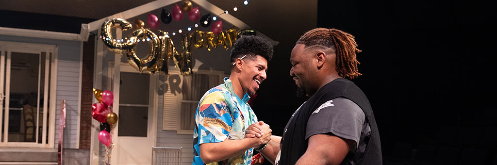 Two people smile and clasp hands in front of a decorated house with balloons and a “Congrats Grad” banner.