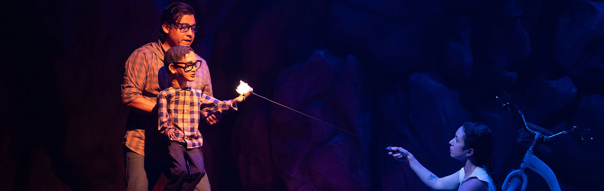 Performer operating a puppet holding a lit stick while another performer kneels nearby, reaching toward the flame.