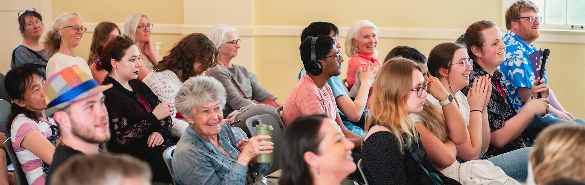 A diverse audience sitting attentively in a room, some smiling and clapping. The group includes people of various ages, and one individual is wearing headphones, while another has a colorful hat.