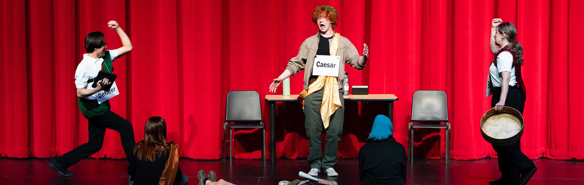 Theatrical performance depicting Julius Caesar’s assassination; student actors on stage with labeled signs, dramatic poses, and Roman-themed costumes.