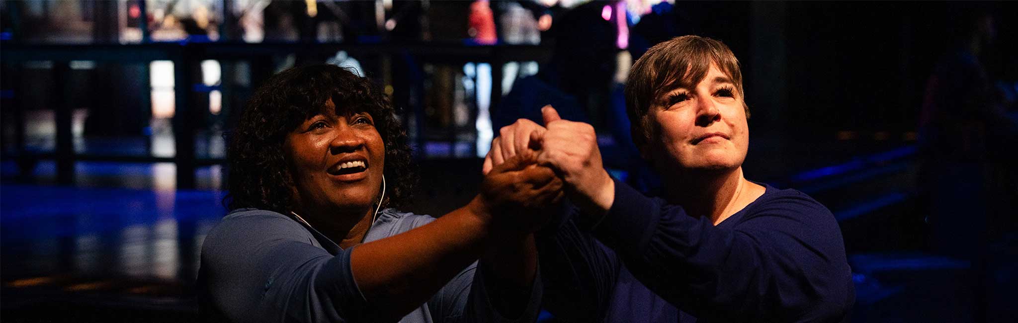 Two people clasp hands and gaze upward with expressions of hope and focus, illuminated by stage lighting in a dark theater setting.